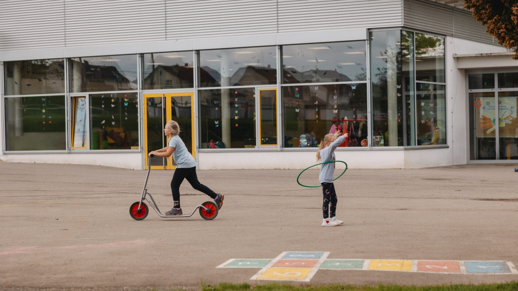 Zwei Schülerinnen spielen im Pausenhof mit Roller und Hoola-Hoop Reifen